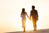 siluety muže a ženy držící se za ruce při chůzi na pláži proti slunci při západu slunce