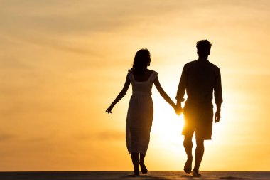 Gün batımında plajda güneşe karşı yürürken el ele tutuşan kadın ve erkeğin siluetleri.