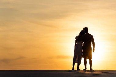 Gün batımında plajda güneşe karşı sarılan kadın ve erkeğin siluetleri.