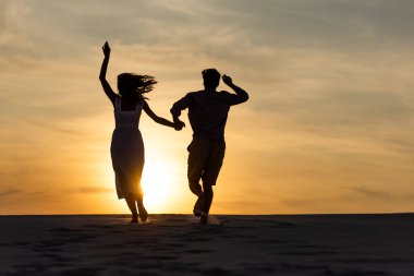 silhouettes of man and woman running on beach against sun during sunset clipart