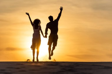 silhouettes of man and woman jumping on beach against sun during sunset clipart