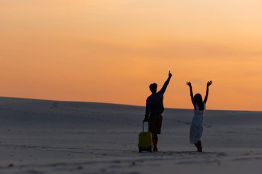 back view of couple walking on beach with hands in air and travel bag at sunset clipart