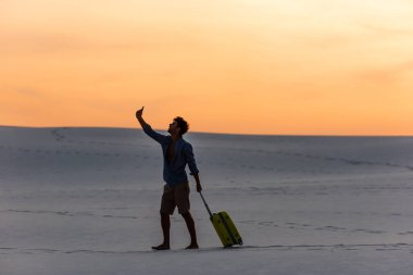 Gün batımında kumsalda seyahat çantası ve akıllı telefonuyla yürüyen bir adamın silueti.