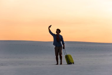 Gün batımında kumsalda seyahat çantası ve akıllı telefonuyla yürüyen bir adamın silueti.