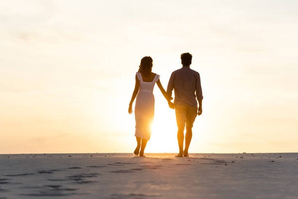 silhouettes of man and woman holding hands while walking on beach against sun during sunset