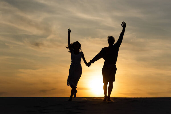 silhouettes of man and woman running on beach against sun during sunset