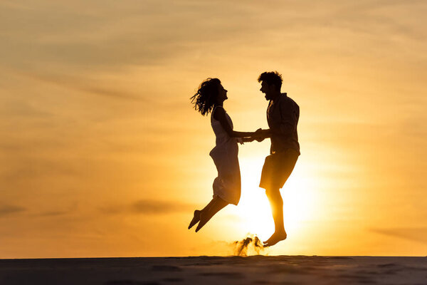 side view of silhouettes of man and woman jumping on beach against sun during sunset