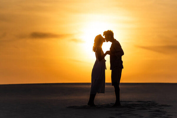 side view of silhouettes of man and woman kissing on beach against sun during sunset