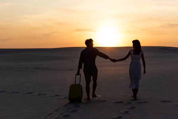 back view of couple walking on beach while holding hands with travel bag at sunset