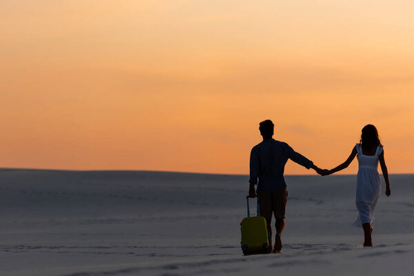 back view of couple walking on beach while holding hands with travel bag at sunset