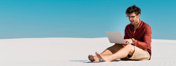 молодой фрилансер на песчаном пляже с помощью ноутбука против ясного голубого неба, панорамный снимок
