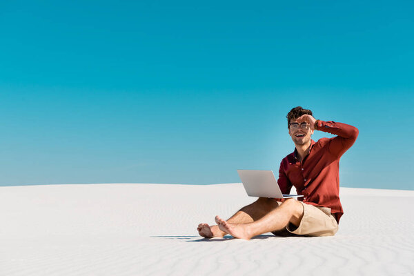счастливый фрилансер на песчаном пляже с ноутбуком на фоне ясного голубого неба
