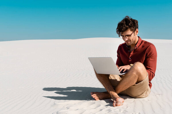 молодой фрилансер на песчаном пляже, используя ноутбук против чистого голубого неба

