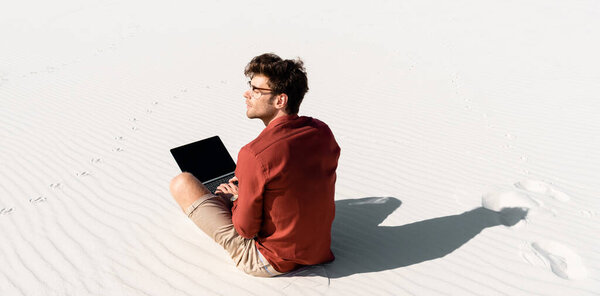 молодой фрилансер сидит на песчаном пляже с ноутбуком
