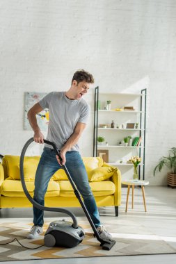 Oturma odasında elektrikli süpürge kullanan ağzı açık yakışıklı bir adam. 