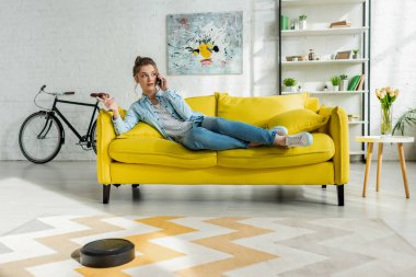 Akıllı telefondan konuşan çekici bir kız robot süpürgesiyle oturma odasında halı yıkıyor. 