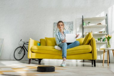 Robot süpürge oturma odasında halı yıkarken dizüstü bilgisayar kullanan çekici bir kadın. 