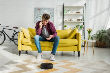 Robot süpürge oturma odasında halı yıkarken akıllı telefondan konuşan neşeli bir adam.