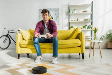 Robot süpürge oturma odasında halı yıkarken akıllı telefondan fotoğraf çeken neşeli bir adam.