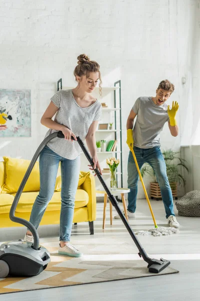 Chica Atractiva Usando Aspiradora Cerca Del Hombre Agitando Mano Mientras — Foto de Stock