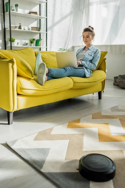 Attraktive Frau Mit Laptop Auf Dem Sofa Während Staubsaugerroboter Teppich — Stockfoto