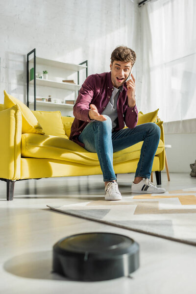 selective focus of happy man talking on smartphone and gesturing near robotic vacuum cleaner washing carpet in living room