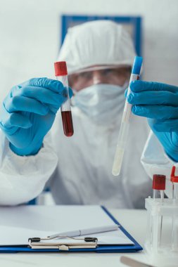 Tehlikeli madde giysileri içindeki biyokimyagerin seçici odak noktası kan örneği ve sıvı içeren test tüpleri tutuyor.
