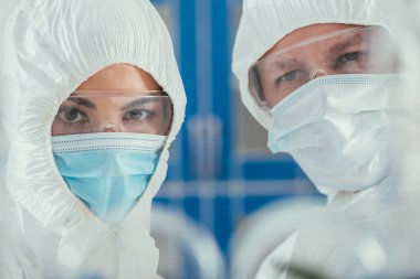Tehlikeli madde odalarındaki biyokimyagerlerin seçici odağı, tıbbi maskeler ve kameralara bakan gözlükler.