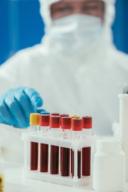Tehlikeli madde giysisi içinde biyokimyacı yakınında kan örnekleri olan test tüplerinin seçici odağı.