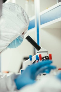 Tehlikeli madde giysileri içindeki biyokimyagerin seçici odağı mikroskoptan bakıyor.
