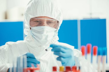 Tehlikeli madde giysisi içindeki biyokimyacı laboratuarda test tüplerinin yanında şırınga tutuyor.