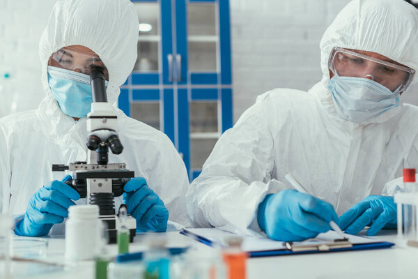 selective focus of biochemist looking through microscope near colleague writing down results on clipboard
