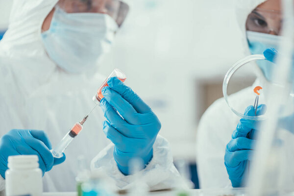selective focus of biochemist taking medicine with syringe near colleague holding petri dish