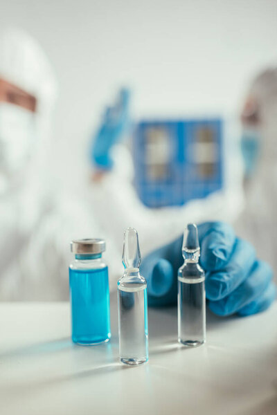 selective focus of biochemist in rubber gloves taking ampule near glass container with blue liquid