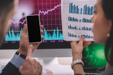 Bilgi güvenliği analistinin seçici odak noktası masadaki çizelgelerle birlikte iş arkadaşının yanındaki akıllı telefona işaret ediyor 