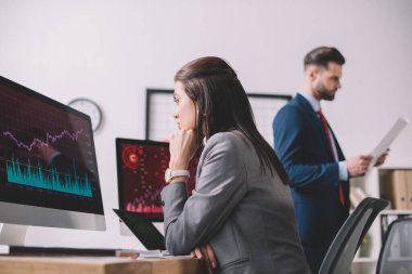 Veri analistinin seçici odak noktası, ofisinde kağıt bulunan meslektaşının yanındaki bilgisayar monitöründeki grafiklere bakıyor. 