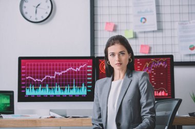 Beautiful data analyst looking at camera near charts on computer monitors in office clipart