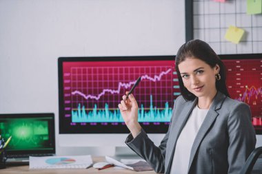Data analyst looking at camera and pointing at charts on computer monitors in office  clipart