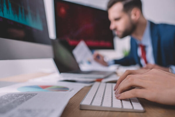 Selective focus of data analyst typing on computer keyboard near colleague at table 