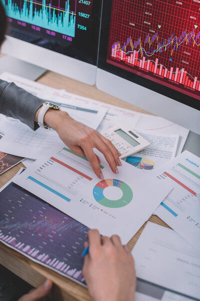 Cropped view of data analysts using charts near calculator and computer monitors on table 