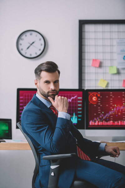 Handsome data analyst looking at camera near charts on computer monitors in office 