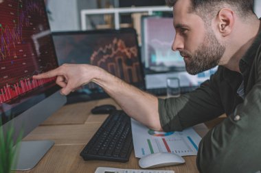 Ofiste çalışırken bilgisayar monitöründeki grafiklere parmak basan bilgi güvenlik analisti yan görünümü 