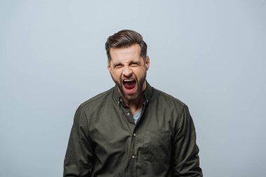 Handsome man screaming at camera isolated on grey clipart