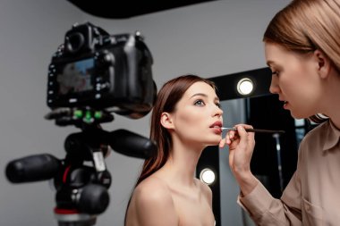 selective focus of makeup artist applying lip gloss on lips of attractive model near digital camera on grey  clipart