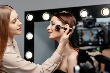selective focus of cheerful makeup artist applying face powder on beautiful model near digital camera  clipart