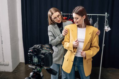 Gözlüklü stilistin seçici odağı dijital kameranın yanındaki mutlu modelde moda cekete dokunuyor 