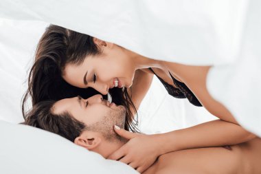 Yatak odasında birbirine sarılan, gülümseyen ve çarşafın altında birbirine bakan çiftler.