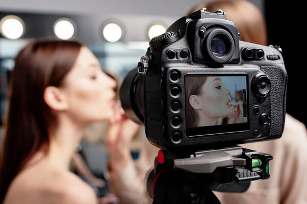 selective focus of digital camera with makeup artist applying lip gloss on beautiful model 