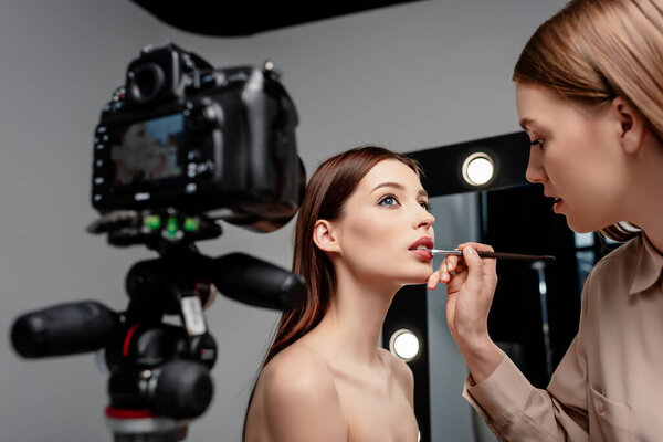 selective focus of makeup artist applying lip gloss on lips of attractive model near digital camera on grey 