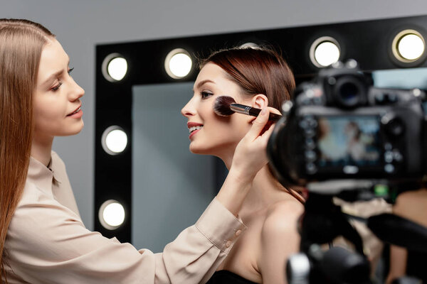 selective focus of cheerful makeup artist applying face powder on beautiful model near digital camera 